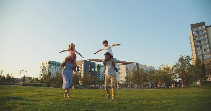 A friendly family of four is relaxing in the city park. They have fun and play. The parents put the children on their shoulders and depict airplanes. The sky is blue above them