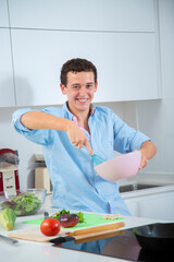 chico joven sonriente bate huevos en un bol rosa con una varilla de cocina azul, en una cocina blanca con verduras y un bol con ensalada de lechuga