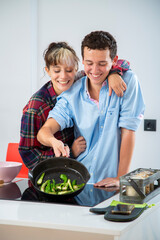 pareja joven sonriente cocinando verduras, pimiento verde, tomate y calabacin, en una cocina blanca con placa de induccion, utilizan un bol rosa, una sarten de hierro y usan herramientas de madera