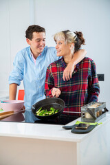 mujer joven sonriente cocina con su pareja pimiento verde, tomate y calabacin, en una cocina blanca con placa de induccion, utilizan un bol rosa, una sarten de hierro y usan herramientas de madera