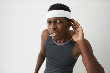 Portrait of African man in sportswear and white headband holding hand over ear frowning, listening...