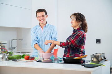 pareja joven sonriente haciendo una masa en un bol rosa, en una cocina blanca con placa de...