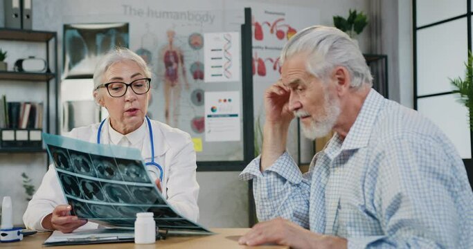 Attending female physician in glasses with stethoscope involved in serious examines the X-ray of the older man and discusses further with his treatment in clinic.