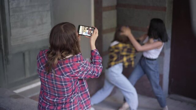 Back view teenage girl filming fight of two angry classmates on smartphone. Caucasian adolescent teenagers fighting outdoors arguing as groupmate recording video for social media