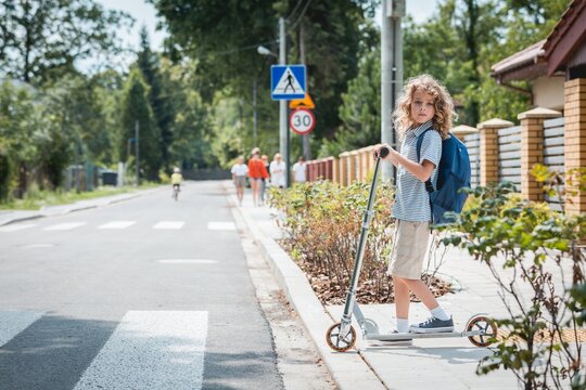 Cute boy with a backpack and scooter is standing in front of a pedestrian crossing on the street