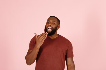 Tired bearded African-American guy model in casual brown t-shirt yawns standing on light pink background in studio closeup