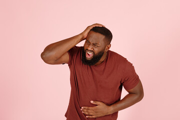 Bearded African-American man patient in t-shirt suffers from severe headache standing on pink background in studio closeup