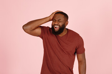 Bearded African-American man patient in t-shirt suffers from severe headache standing on pink background in studio closeup