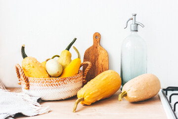 Wooden kitchen table with colorful pumpkins in knitted basket, carving board, and water bottle. Autumnal orange, yellow colors, blanket and gas stove