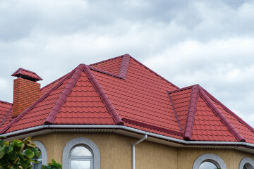 A roof made of red metal tiles with a chimney against a cloudy sky.