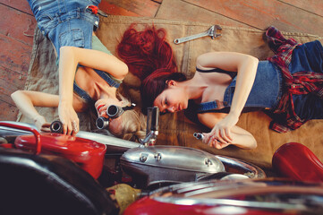 Plakat Young beautiful female mechanic working on a custom retro motorcycle. A talented girl in a blue overalls. She uses a ratchet wrench to tighten the nut bolts. Creative Authentic Workshop Garage