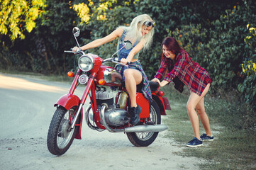 Obraz na płótnie Canvas Young beautiful women ride a retro motorcycle. A talented girl in a blue overalls. They push the motorcycle, the merry go to the road. sale of spare parts. Place for text
