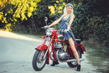 Naklejka na ściany i meble The biker is a beautiful cheerful girl sitting on her motorcycle, a little crazy. Wear short shorts and glasses. holding the steering wheel the wind blows the hair. Looks into the distance at the road