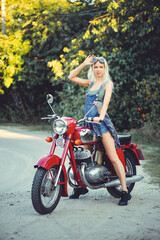 Obraz na płótnie Canvas The biker is a beautiful cheerful girl sitting on her motorcycle, a little crazy. Wear short shorts and glasses. holding the steering wheel the wind blows the hair. Looks into the distance at the road