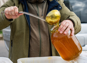 The man's cut-off hands are pouring a mass of bee honey into a glass jar using a ladle. Agricultural Products Fair.
