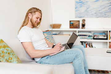 Bearded blonde man working with laptop while sitting on couch