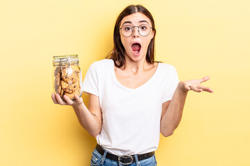 young hispanic woman amazed, shocked and astonished with an unbelievable surprise. cookies bottle...