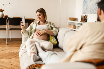 Pregnant woman taking selfie on cellphone while sitting at couch