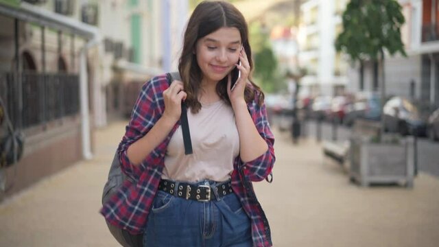Front view of charming confident Caucasian teenage girl talking on phone smiling walking on city street. Beautiful college student strolling with backpack outdoors chatting. Confidence and lifestyle