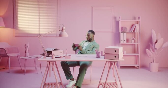 African man playing smartphone game app in pink pastel room studio monocolor creative office with monochrome design. Using laptop happy wth the results Creative procrastination alone in white cloth