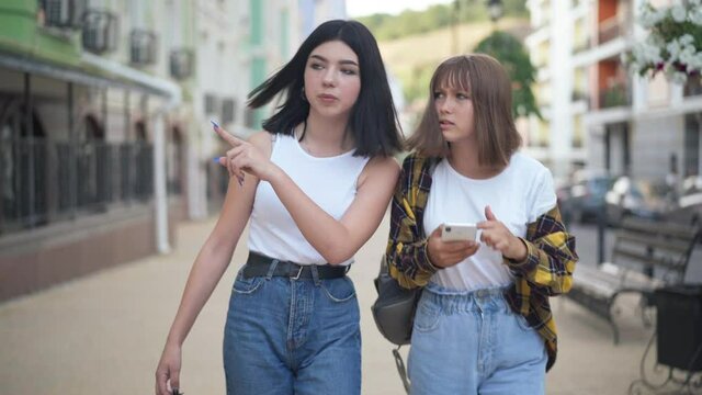Teenage Caucasian girls talking walking in European city searching address with online map in smartphone. Portrait of positive charming teenagers strolling outdoors travelling together