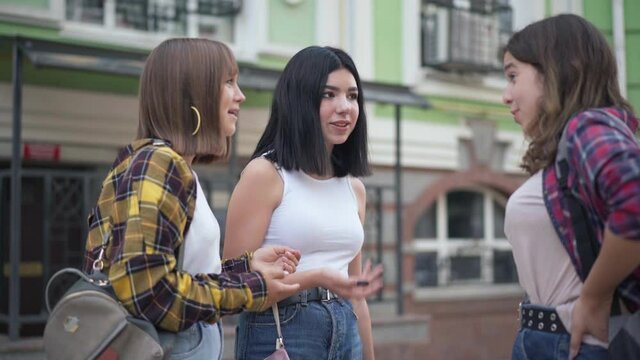 Positive Caucasian teenage girls standing on city street talking laughing. Three charming confident teenagers gossiping outdoors meeting after college. Rumors and lifestyle concept