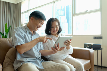 Happy elderly Asian couple watching funny photos or videos online on digital tablet , aged couple...