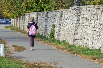 Fototapeta na wymiar A girl with a backpack walks along a path along a stone wall on an autumn day