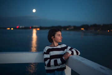 Fototapeta premium Portrait of preteen girl looking aside outdoors on pier by sea at dusk, holiday concept.