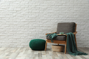 Stylish knitted pouf and comfortable armchair near white brick wall indoors, space for text....