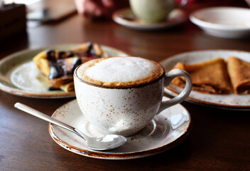 Coffee in a beautiful ceramic cup on a wooden table in a cafe on the background of plates with pancakes. Cappuccino coffee.