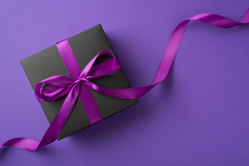 Top view photo of stylish black giftbox with violet satin ribbon bow on isolated violet background with blank space