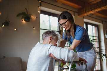 Healthcare worker or caregiver visiting senior woman indoors at home, helping her to walk.