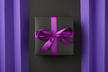 Top view photo of black giftbox with purple ribbon bow on isolated back and violet sheet with vertical folds background
