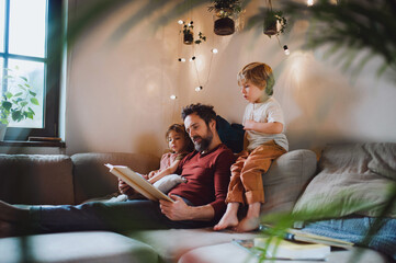 Mature father with two small children resting indoors at home, looking at photo album. - obrazy, fototapety, plakaty