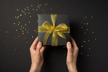 First person top view photo of girl's hands giving black giftbox with golden mesh ribbon bow over...