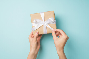 First person top view photo of hands unpacking craft paper giftbox with white ribbon bow on isolated pastel blue background