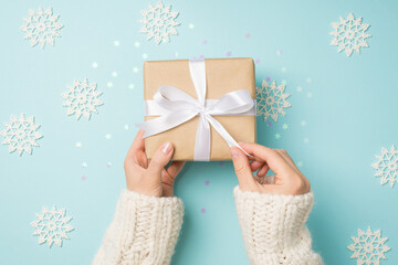 First person top view photo of hands in white sweater unpacking craft paper giftbox with white ribbon bow over big decorative snowflakes and shiny confetti on isolated pastel blue background