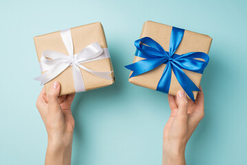 First person top view photo of hands holding two craft paper gift boxes with vivid blue and white ribbon bow on isolated pastel blue background