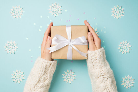 First person top view photo of girl's hands in white sweater holding craft paper giftbox with white ribbon bow over big decorative snowflakes and shiny sequins on isolated pastel blue background