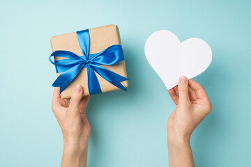 First person top view photo of hands holding craft paper giftbox with blue satin ribbon bow and white paper heart on isolated pastel blue background with blank space