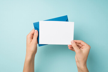 First person top view photo of hands holding blue envelope and white card on isolated pastel blue background with empty space