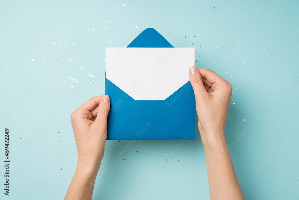 Wall mural First person top view photo of hands holding open blue envelope with white card over sequins on isolated pastel blue background with empty space