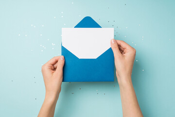 First person top view photo of hands holding open blue envelope with white card over sequins on isolated pastel blue background with empty space