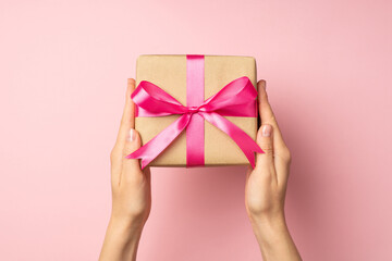 First person top view photo of hands holding stylish craft paper giftbox with pink ribbon bow on isolated pastel pink background