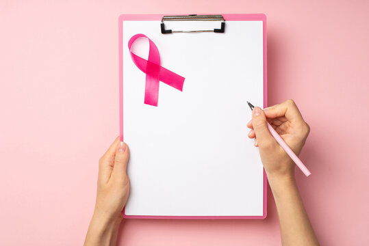 First person top view photo of hands holding pink pen and pink clipboard with paper sheet and pink ribbon symbol of breast cancer awareness on isolated pastel pink background with empty space