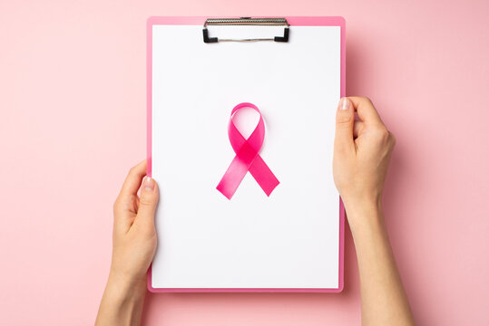 First person top view photo of woman's hands holding pink clipboard with paper sheet and pink ribbon symbol of breast cancer awareness in the middle on isolated pastel pink background