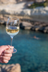 Man's hand holding glass of white dry white wine with view on rocks and blue sea bay water near Protaras touristic town on Cyprus