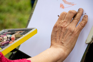 Street artist holding a box with multicolored crayons and pencils for drawing.Close up photography of hands and drawing supplies.He drawing behind the easel with white paper.