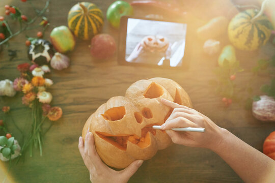 Halloween concept: woman carves a mouth in a pumpkin to make a jack o'lantern on a wooden table with vegetables using video manual on tablet. Top view. High quality image